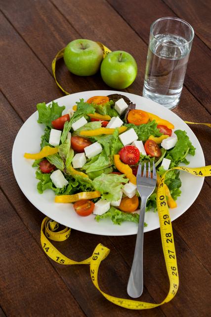 Image showing a plate of fresh salad with feta cheese, cherry tomatoes, and yellow bell peppers, accompanied by two green apples, a glass of water, and a measuring tape. Ideal for illustrating concepts related to diet, weight loss, healthy eating, and fitness. Perfect for use in health articles, diet plans, wellness blogs, and fitness-focused websites.