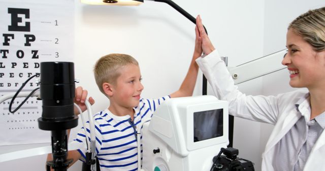 A young boy is giving a high-five to an optometrist in an eye clinic. There is an eye chart visible in the background, and the boy is seated at eye testing equipment. Perfect for use in health care articles, pediatric health features, vision care advertisements, or ophthalmology clinic promotions.