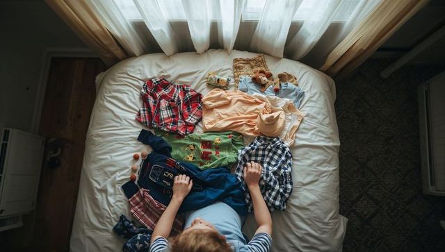 Child organizing colorful kids clothing and toys on bed under soft daylight window for packing