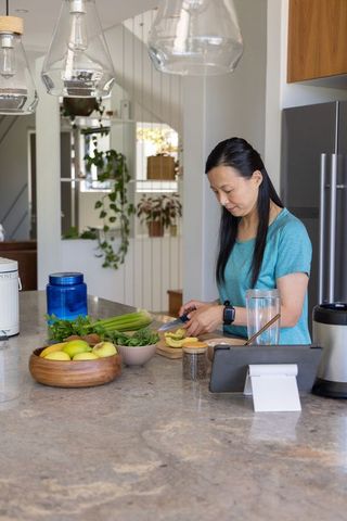 Mature asian woman preparing avocado on kitchen island with tablet and fresh produce