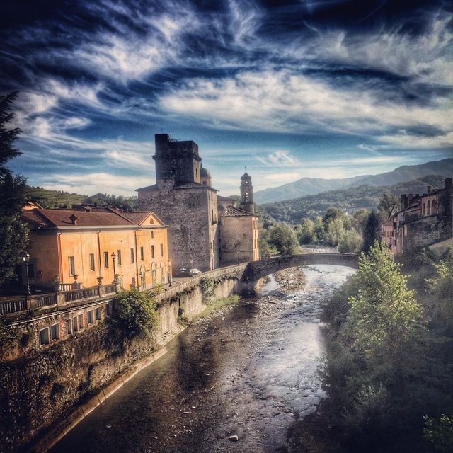 Depicts a picturesque Italian village with historical castle by a river, featuring a stone bridge and towering ancient buildings under a dramatic sky and surrounded by rolling hills. Useful for travel brochures, historical documentaries, promotional material for tourism, or articles about Mediterranean destinations.