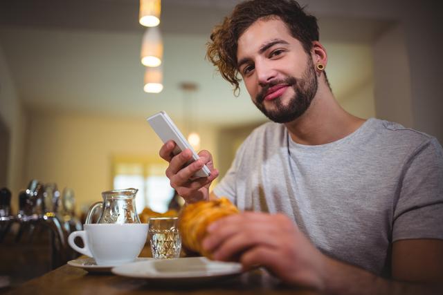 Young man enjoying breakfast in a cozy café, using his mobile phone while holding a croissant. Ideal for lifestyle blogs, technology advertisements, and social media content promoting casual dining or modern communication.