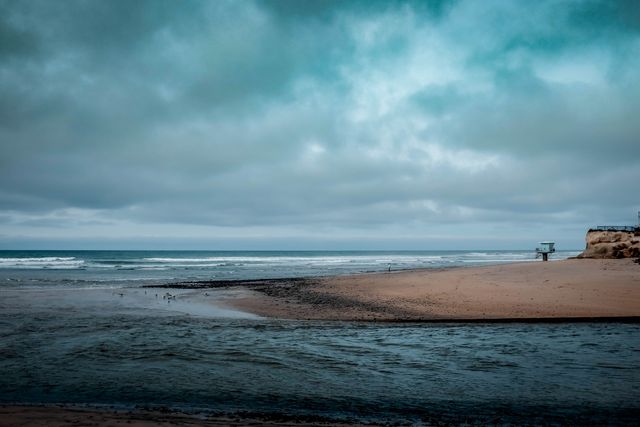 Overcast sky casting a dramatic mood over a tranquil beach with gentle waves. Sand and water meet, creating a peaceful shoreline view. Ideal for backgrounds, travel promotions, environmental articles, or relaxation-themed content.