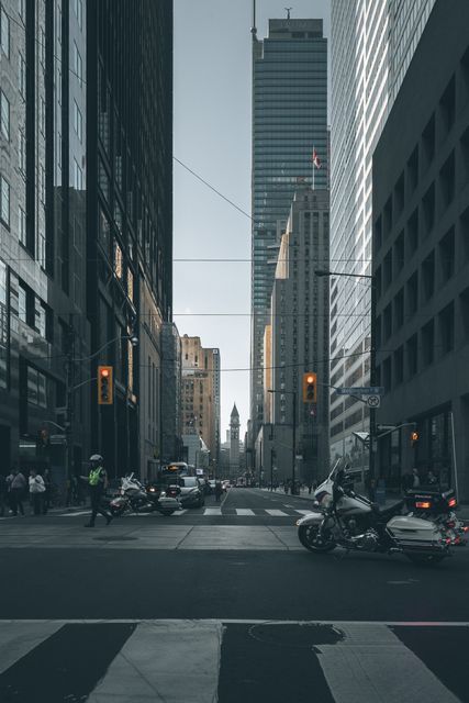Image depicting a city street flanked by tall skyscrapers, with a crosswalk in the foreground and traffic, including motorcycles, present. This type of image is suitable for use in articles, blogs, and websites focusing on urban living, architecture, transportation, and city planning.
