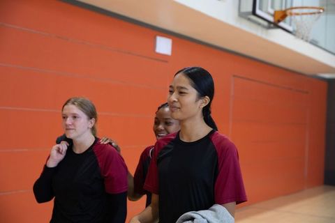 Multiracial high school girls walking in gym wearing maroon jerseys holding sweatshirt