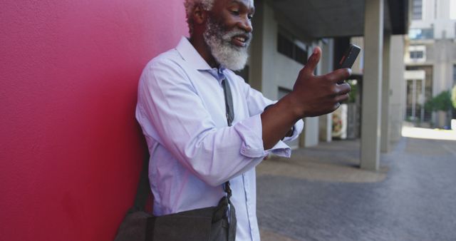 Elderly man standing against a red wall, using a smartphone in an outdoor urban environment. This can be used for themes involving technology use among seniors, urban lifestyles, or independent living for older adults.