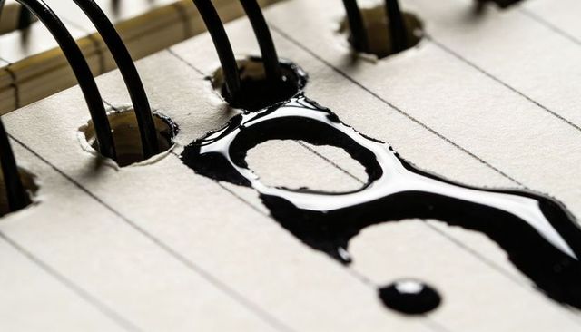 Glossy black ink spilling and spreading on lined notebook near spiral binding, macro