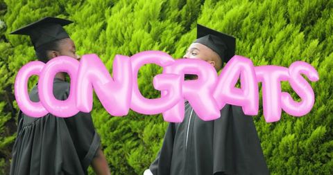 Graduates celebrating with pink congrats balloon wearing gowns and mortarboards outdoors