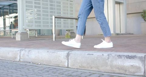 Casual urban walking on curb with white sneakers and cropped blue jeans, sidewalk lifestyle