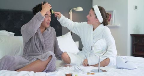 Diverse couple sharing cozy skincare routine in plush robes, woman applying facial brush