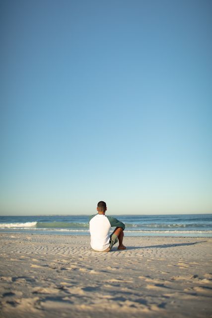 Man sitting on sandy beach facing the ocean under a clear blue sky. Ideal for themes of relaxation, solitude, and nature. Perfect for travel brochures, wellness blogs, and inspirational content.
