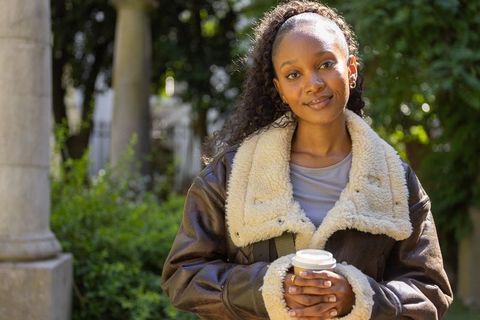 African woman holding coffee cup and smiling in sunlit courtyard wearing shearling jacket