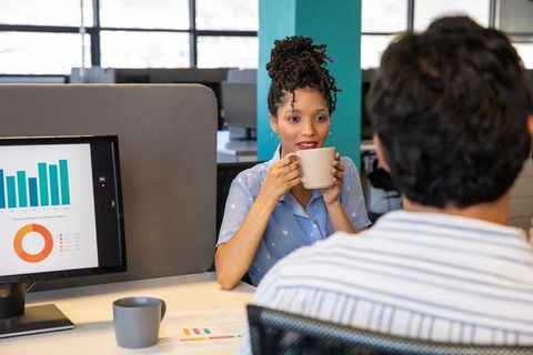 Smiling woman drinking coffee and discussing charts with coworker in open-plan office