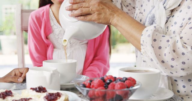 Family member pouring tea while having breakfast with fresh berries outdoors. Ideal for themes like family bonding, morning routines, healthy lifestyle, and summer activities. Can be used for advertisements, blog posts, and social media content focusing on wellness and family time.