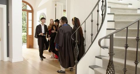 Diverse graduates gathering in home foyer wearing gowns and mortarboards near staircase