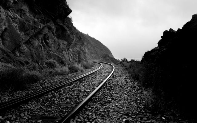 Tracks curving through rocky terrain in dramatic black-and-white composition conveying themes of travel, journey, and adventure. Suited for concepts of wanderlust, perseverance, and exploration. Great for travel blogs, motivational posters, or transport industry's promotional materials.