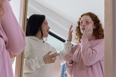 Diverse friends applying heart-shaped under-eye patches at mirror for morning self-care