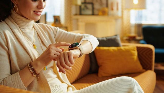 Smiling woman checking smartwatch on wrist while relaxing on mustard sofa wearing turtleneck