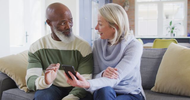 Happy senior diverse couple in living room sitting on sofa, using smartphone, making image call. retirement lifestyle, at home with technology.