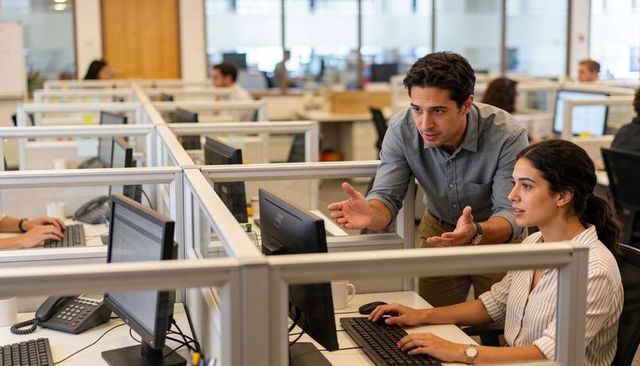 Male mentor explaining to female colleague while she types at computer in modern open-plan office