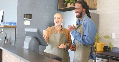Diverse baristas laughing and pointing at touchscreen register in modern coffee shop