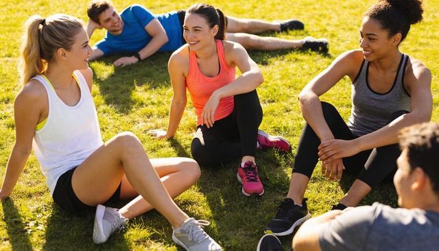 Group of adults relaxing and chatting after outdoor workout on sunny park lawn