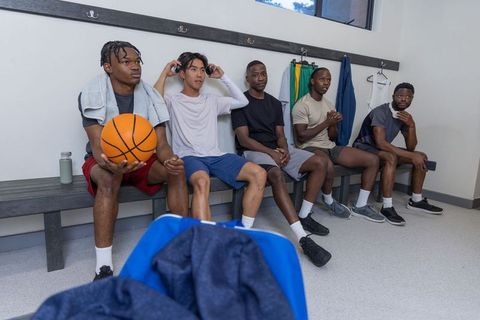 Diverse basketball players sitting on locker bench holding ball and wearing headphones