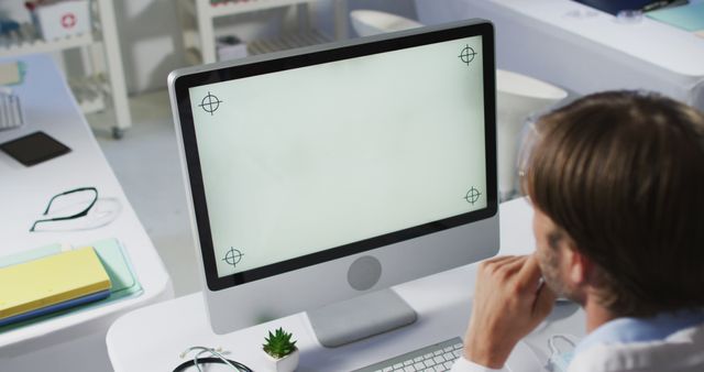 Man sitting at desk, looking at computer with blank white screen featuring calibration markers. Suitable for features on technology use in business settings, professional work environments, or discussions on computer troubleshooting and office productivity.