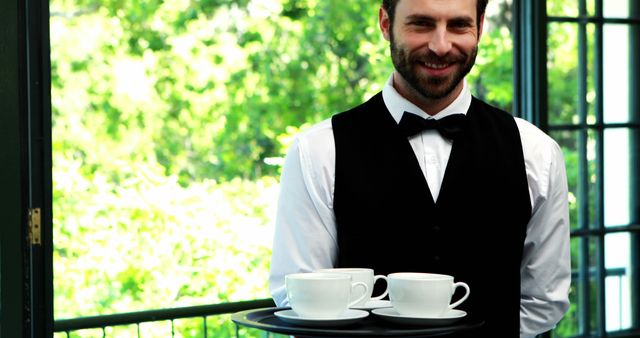 Smiling waiter holding a tray with coffee cups, dressed in a white shirt, black vest, and bow tie. Bright outdoor setting in a cafe with lush greenery visible through the windows. Ideal for depicting hospitality, customer service, restaurant environment, or promotional content for cafes and coffee shops.