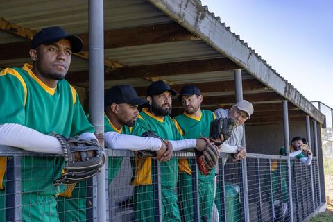 Diverse baseball teammates leaning in dugout wearing green uniforms and gripping gloves