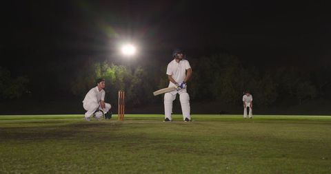 Floodlit night cricket action with batsman, wicket-keeper and fielders on grass pitch