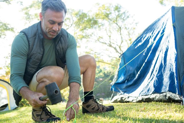 Man setting up a tent at a campsite, preparing for a camping trip. Ideal for use in articles or advertisements related to outdoor activities, camping gear, travel, and adventure. Can also be used in blogs or social media posts promoting nature excursions and leisure activities.