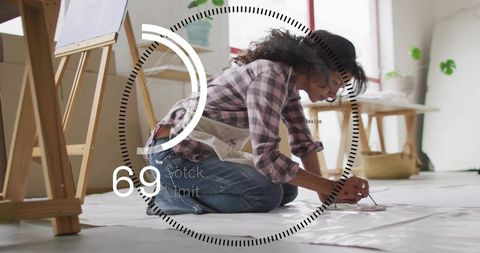 Kneeling artist painting on drop cloth in sunlit loft studio wearing plaid shirt and apron