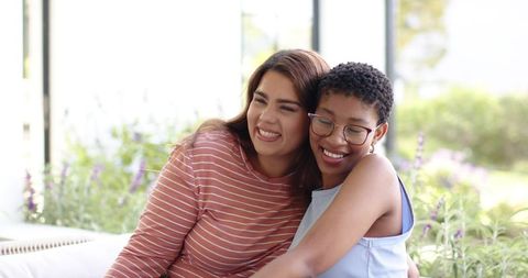 Sunlit friends hugging on sofa with plants and eyeglasses, cozy home lifestyle portrait