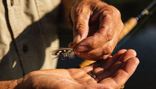 Seasoned angler hand-tying feather fly over palm with weathered hands and fishing rod