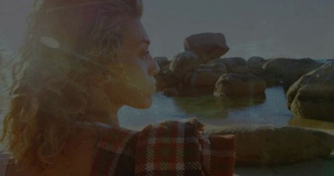 Woman gazing over rocky shoreline wrapped in red plaid blanket at golden hour