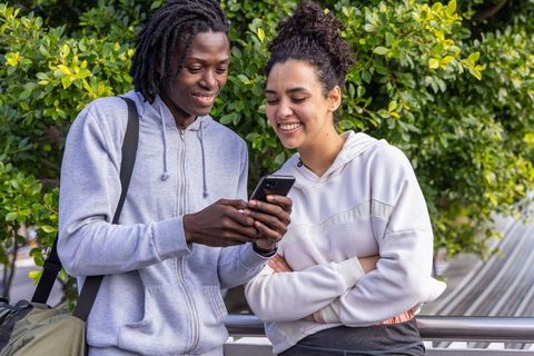 Smiling multicultural young adults sharing smartphone outdoors near urban greenery
