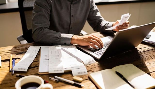 Freelancer organizing receipts and doing bookkeeping on laptop at rustic wooden desk with coffee