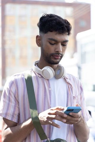 Indian man using smartphone while wearing headphones and pink striped shirt outdoors