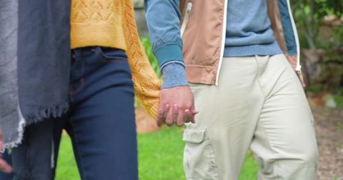 Diverse couple walking hand in hand in park wearing mustard knit and tan jacket