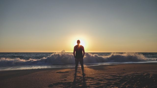 Person standing on sandy beach facing ocean during sunset with waves crashing. Ideal for travel blogs, inspirational quotes, relaxation themes, or promoting beach vacations.