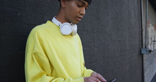 A teenage boy wearing a bright yellow sweatshirt and headphones around his neck is using a smartphone outside on a street. This image is suitable for use in articles or advertisements focusing on teenage fashion, technology usage, outdoor activities, and street culture.