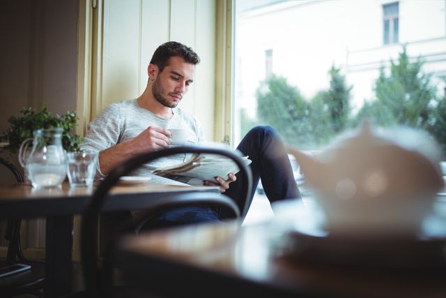 Young man sitting in a cozy café, enjoying a cup of coffee while reading a newspaper. Ideal for use in lifestyle blogs, coffee shop advertisements, or articles about relaxation and leisure activities.