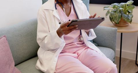 African nurse using tablet on clinic sofa wearing pink scrubs and white lab coat