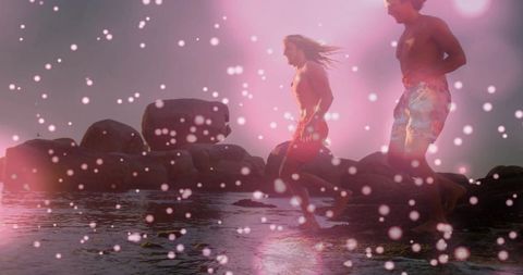 Three friends running along rocky shoreline at dusk with pink bokeh and lens flare