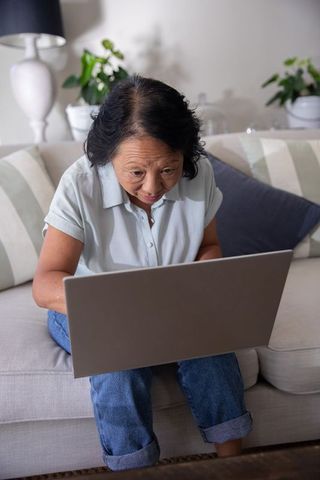 Senior asian woman leaning forward on sofa using laptop for online learning and calls