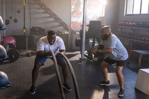 African man swinging heavy battle ropes intense gym training with coach for strength