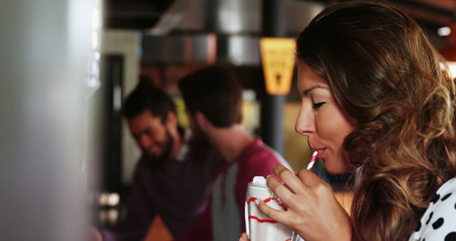 Young woman savoring a milkshake in a lively cafe, with two friends chatting in the background. Ideal for use in advertising campaigns related to food and beverages, casual dining, social gatherings, or lifestyle articles.