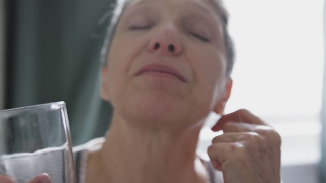 A senior Caucasian woman drinks water and swallows medication while sitting on her bed. This image conveys themes of health awareness, daily life routines for older adults, and the importance of staying hydrated. It can be used for advertising healthcare products, senior living facilities, medical articles, and wellness campaigns.