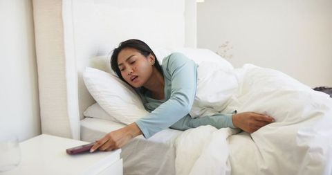 Mid-adult asian woman reaching for smartphone from cozy bed in bright minimalist bedroom